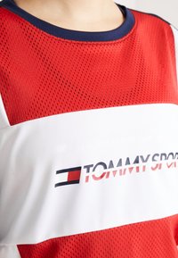 Zbliżenie na czerwoną i białą sportową koszulkę z siateczki z granatowym kołnierzykiem i logo "Tommy Sport" na klatce piersiowej.