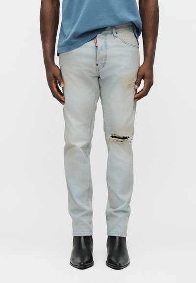 Jean déchiré bleu clair porté par une personne vêtue d'une chemise bleue et de bottines noires, visible de la mi-torse jusqu'aux pieds, sur un fond blanc.