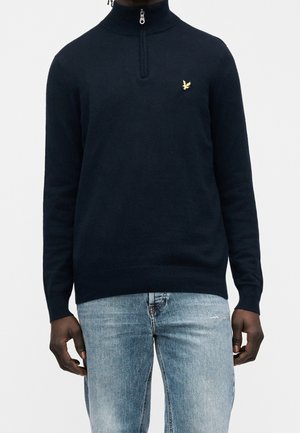 Mørkhudet person iført en mørkeblå kvart-lynlåstrøje med et lille gyldent fuglelogo og lyseblå jeans.