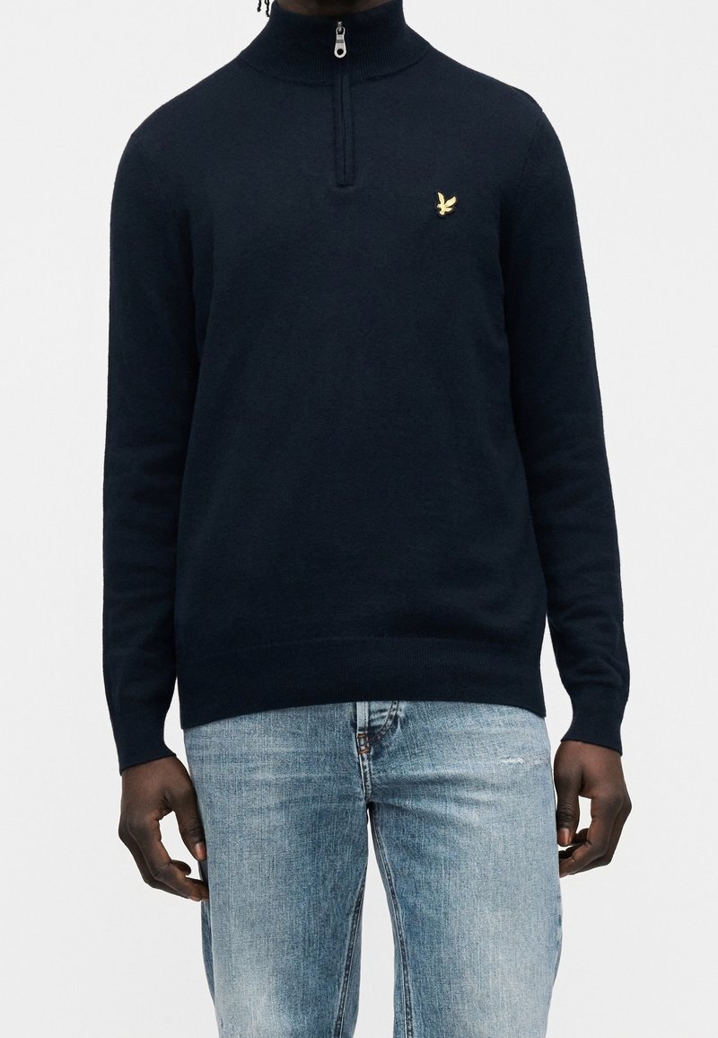 Personne à la peau foncée portant un pull bleu marine à fermeture éclair au quart avec un petit logo d'oiseau doré et un jean bleu clair.