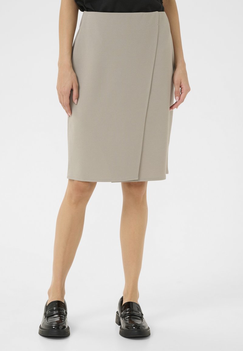 Beige kokerrok tot de knie met een wikkeldesign, gemaakt van een soepele stof. Draagt met zwarte gepatineerde loafers. Eenvoudige, strakke lijnen.