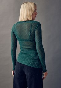 Grøn langærmet top lavet af gennemsigtigt stof med glitrende detaljer. Har et tætsiddende design og rynket ryg. Kombineret med mørke jeans.