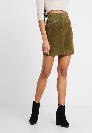 Kobieta w oliwkowej zamszowej mini spódnicy z przednimi kieszeniami, w połączeniu z beżowym krótkim topem i czarnymi, wysokimi botkami na obcasie z zamkami.