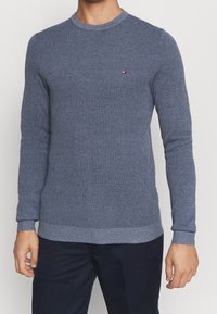 Miestny modrý textúrovaný pletený sveter s malým logom Tommy Hilfiger na ľavej hrudi, v kombinácii s tmavomodrými nohavicami.