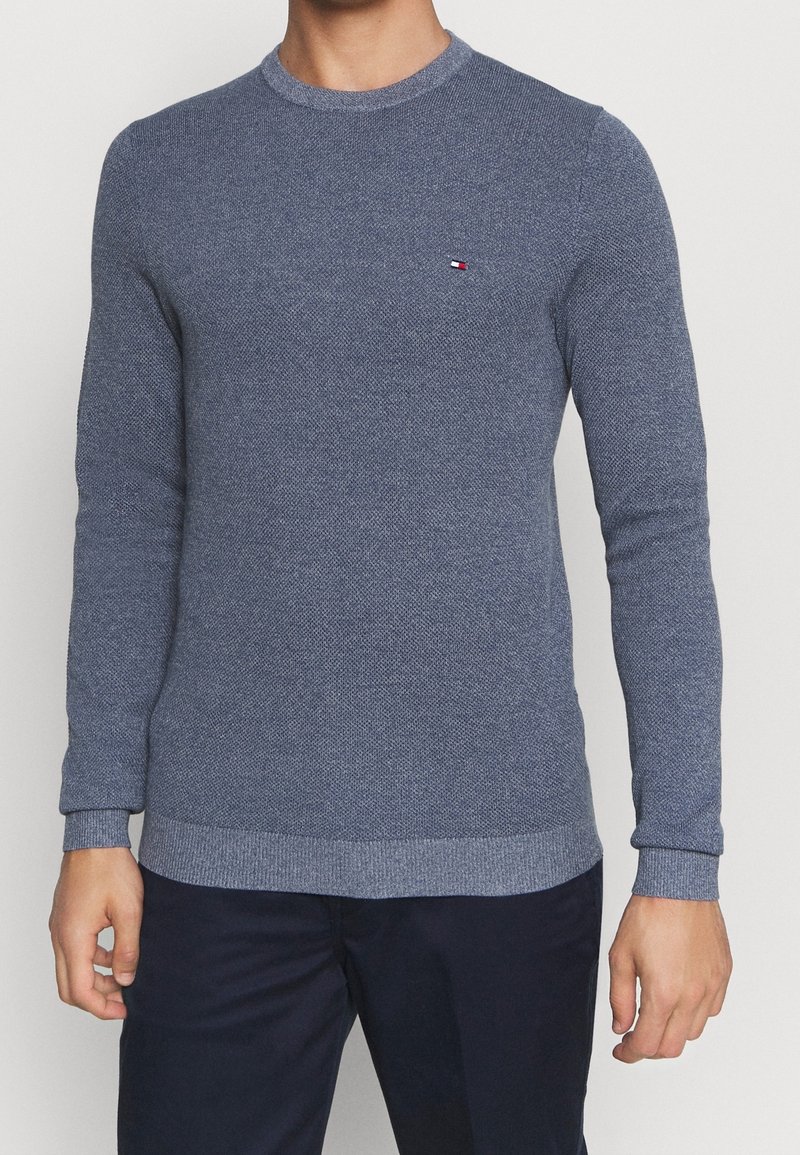 Miestny modrý textúrovaný pletený sveter s malým logom Tommy Hilfiger na ľavej hrudi, v kombinácii s tmavomodrými nohavicami.