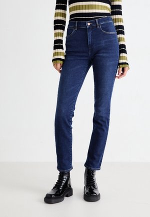 Osoba ubrana w ciemnoniebieskie obcisłe dżinsy, czarne sznurowane botki sięgające kostki oraz cienki sweter z długim rękawem w paski w kolorach czarnym, białym i oliwkowo-zielonym.