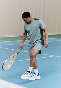 Atleta com um traje atlético verde claro com detalhes em preto, segurando uma raquete de ténis, em pé num court azul com uma bola de ténis amarela.