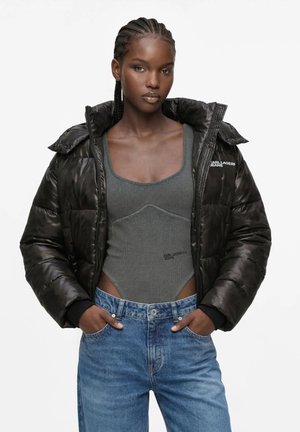 Kvinde med flettet hår iført en sort dunjakke, grå tætsiddende bodysuit og blå jeans, stående med hænderne i lommerne mod en hvid baggrund.