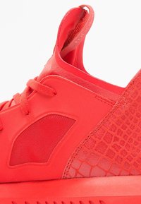 Červená atletická bota s texturovanou boční částí připomínající plazí kůži, hladkým svrchním materiálem a tónovacími tkaničkami. Minimalistický design s poutkem.