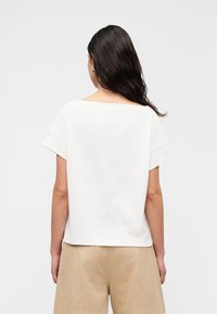 Λευκό τοπ με κοντά μανίκια και φαρδύ ντεκολτέ, με απαλό ύφασμα, άνετη εφαρμογή, εμφανίζεται σε συνδυασμό με χακί σορτς, πίσω όψη.