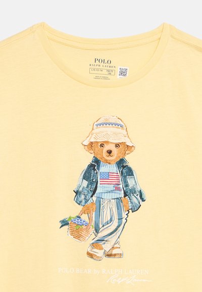 Κίτρινο πουκάμισο με το Polo Bear που φοράει καπέλο τύπου bucket, τζιν μπουφάν, ριγέ παντελόνια και κρατάει ένα καλάθι με σταφύλια.