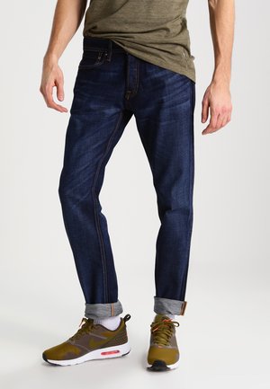Donkerblauwe spijkerbroek met een slim fit, zichtbare stiksels en opgerolde boorden, gecombineerd met olijfgroene sportschoenen met witte accenten.