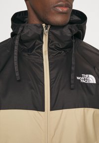 Personne portant une veste à capuche North Face beige et noire avec une fermeture éclair beige et un logo blanc sur la poitrine.