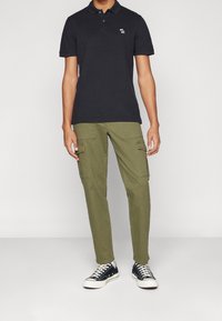 Černé polo tričko s krátkým rukávem, vyšitým logem na hrudi, spárované s olivově zelenými cargo kalhotami s bočními kapsami a černými teniskami.