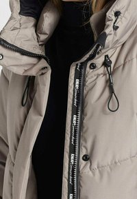 Beige gesteppte Pufferjacke mit hohem Kragen, ausgestattet mit einem schwarzen Reißverschluss, Logo-Band entlang des Reißverschlusses und elastischen Zugbändern zur Anpassung.