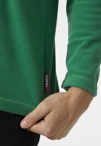 Zelený fleecový svetr s měkkou strukturou, s prošitým lemem a viditelnou nášivkou Polartec® na boku, ruka držící látku.