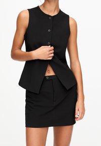 Blouse noire sans manches avec un col montant, des boutons sur le devant et une coupe ajustée, associée à une jupe noire assortie et cintrée.