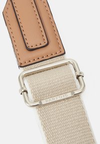 Canvasrem i beige med brunt läderaccent. Har ett spänne i silvermetall, präglad logga och kontrasterande sömmar.