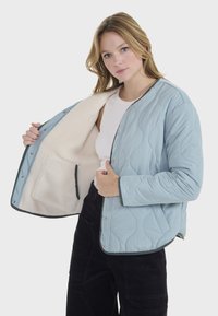Chaqueta acolchada de color azul claro con un suave forro color crema, que cuenta con botones a presión, bolsillos laterales y un ribete oscuro contrastante a lo largo de los bordes.