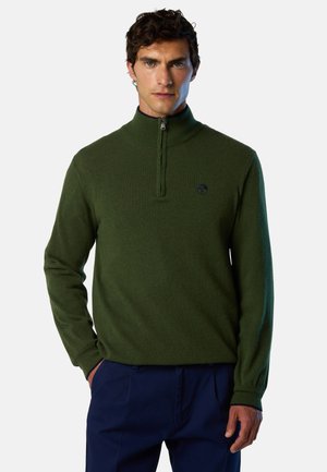 Hombre con un suéter de media cremallera verde oscuro con un pequeño logo en el pecho, combinado con pantalones azul marino, posando frente a un fondo blanco.