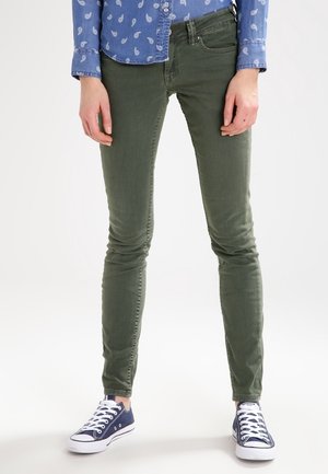 Personne portant un pantalon slim vert olive, une chemise à motifs bleue à boutons et des baskets basses bleu marine à lacets blancs.