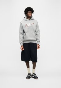 Γκρι φούτερ με κουκούλα με κόκκινο λογότυπο Nike, μαύρη βερμούδα, μπεζ κάλτσες και μαύρα αθλητικά παπούτσια. Τα υλικά φαίνονται απαλά με άνετη εφαρμογή.