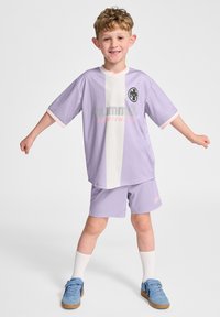 Lila sporttröja och shorts-set med vit panel, rosa detaljer, logotyp på bröstet, korta ärmar och avslappad passform. Bärs med blå skor och vita strumpor.