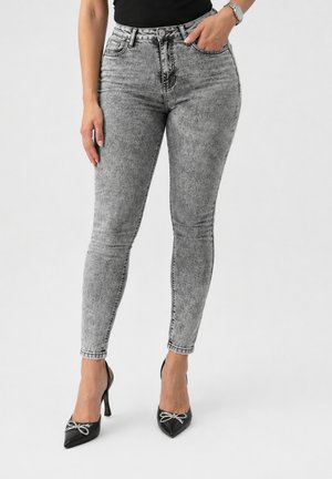 Frau trägt graue Skinny-Jeans mit hohem Bund und Acid-Wash und schwarze spitze High Heels mit Schleifendetail, steht vor weißem Hintergrund.