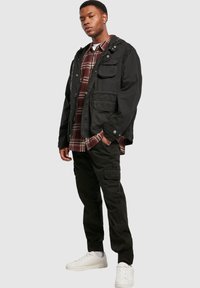 Black hooded jacket with multiple pockets, worn over a red and white plaid shirt, paired with black cargo pants and white sneakers.