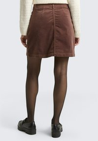 Bruine ribfluwelen rok met een rechte snit, achterzakken en een afgewerkte zoom. Gecombineerd met zwarte panty’s en robuuste zwarte schoenen.