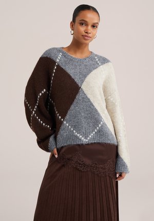 Femme portant un pull color-block marron, gris et crème avec un motif losange, associé à une jupe plissée marron avec un bord en dentelle.