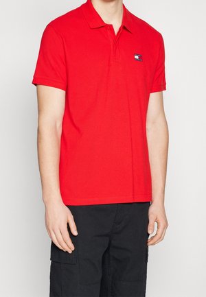 Red polo shirt with a classic collar, short sleeves, and a small navy logo patch on the left chest. Fabric appears smooth and breathable.
