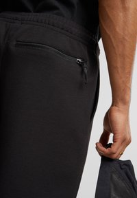 Pantalones deportivos negros con un bolsillo con cremallera. La tela parece suave y tiene una textura lisa. Una mano sostiene una prenda negra, mostrando un detalle de malla.