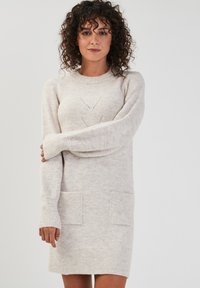 Robe pull tricotée beige clair avec des poignets côtelés, deux poches avant et un col rond. Présente des motifs en diamant subtils sur la poitrine.