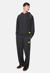 Czarna bluza z kapturem i pasujące dresy z miękkiego materiału, z zielonymi przeszyciami i kwadratowymi metkami logo. Strój ma luźny krój.