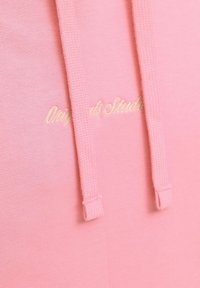Hoodie rosa con tessuto testurizzato, caratterizzato da cordini e logo ricamato in giallo chiaro. Messa a fuoco sull'aspetto casual e morbido.