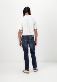 Camisa blanca de manga corta con un diseño texturizado, combinada con jeans azul oscuro que tienen bolsillos traseros y un parche de cuero. Se lleva con zapatillas de deporte con un patrón.