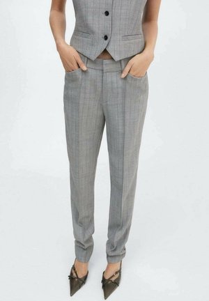 Vrouw draagt grijze pantalon met krijtstrepen en bijpassend waistcoat, handen in de zakken, gecombineerd met puntige zwarte hakken.