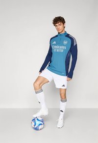 Zils un tumši zils garām piedurknēm sporta tops ar "Emirates Fly Better" logotipu, baltas šortas un zeķes ar melniem svītrām, stāvot uz krāsainas futbola bumbas.