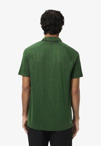 Camisetta verde a maniche corte con un motivo geometrico, colletto e texture liscia. Il design sul retro presenta un marchio sottile vicino al colletto.
