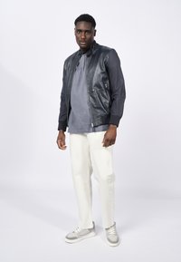 Chaqueta bomber de cuero con mangas en azul marino oscuro, camiseta gris, pantalones blancos y zapatillas grises con detalles de acento. Textura suave, diseño ajustado.