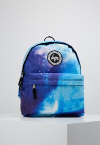 Reppu, jossa on galaksikuvio sinisissä ja purppura sävyissä, mustalla logo-ompeluksella ja vetskarella varustettu etutasku. Valmistettu kestävästä kankaasta.