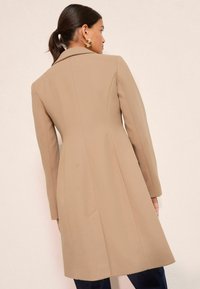 Béžový prispôsobený kabát so štruktúrovaným golierom, dlhými rukávmi a bočnými vreckami. Hladká textúra s priliehavým strihom a detailmi ševmi na zadnej strane.