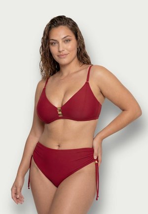 Femme aux cheveux bruns ondulés portant un maillot de bain deux-pièces rouge, debout avec une main sur la hanche sur un fond uni.