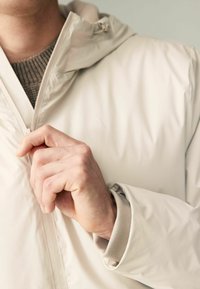 Jachetă impermeabilă de culoare bej deschis, cu glugă, dotată cu fermoar bidirecțional, manșete cu nervuri și o textură mată, netedă.