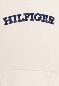 Felpa crema con ricamo in blu della scritta "HILFIGER" ad arco. Il tessuto appare morbido e con una texture liscia; presenta una tasca frontale.