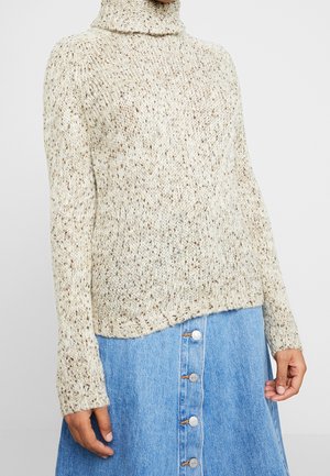 Femme portant un pull à col roulé crème tacheté et une jupe en jean bleue à boutons devant, mains relâchées le long du corps.
