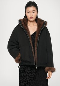 Giacca nera imbottita con fodera e polsini in pelliccia sintetica marrone, dotata di chiusura con zip e cappuccio oversize. Indossata sopra un vestito nero a trama.