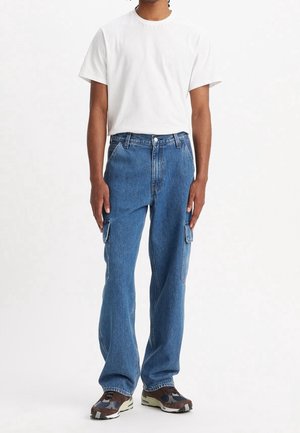 Personne portant un t-shirt blanc uni, un jean cargo bleu avec des poches latérales et des baskets marron et noires, debout devant un fond blanc.