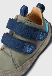 Grüner Lederschuh mit navyblauen Klettverschlüssen. Weicher Wildlederbesatz und ein kleines blaues Bärenlogo am Riemen. Abgerundete Form mit einer Fersenschlaufe.